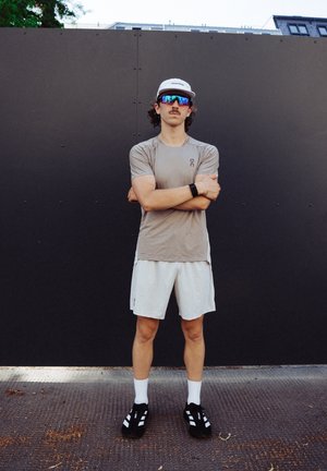 Conjunto atlético que incluye una camiseta de manga corta marrón claro, pantalones cortos beige, zapatos negros y una gorra blanca con gafas de sol con espejo.