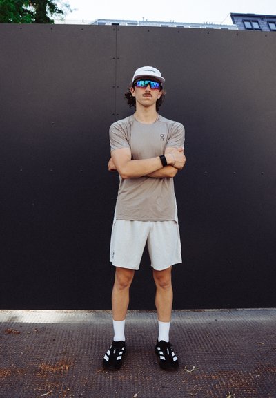 Conjunto atlético que incluye una camiseta de manga corta marrón claro, pantalones cortos beige, zapatos negros y una gorra blanca con gafas de sol con espejo.