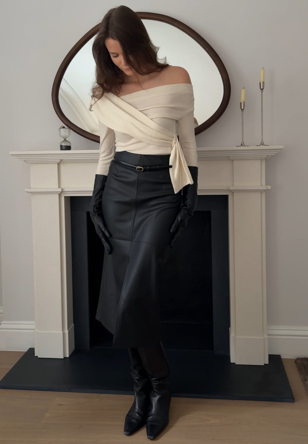 Γυναίκα που φοράει μπεζ τοπ με έξω τους ώμους, μαύρη δερμάτινη φούστα, μαύρα γάντια και μπότες, στέκεται μπροστά σε ένα τζάκι με οβάλ καθρέφτη και κεριά.