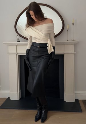 Mujer con blusa beige de hombros descubiertos, falda de cuero negra, guantes y botas negras, de pie frente a una chimenea con un espejo ovalado y velas.