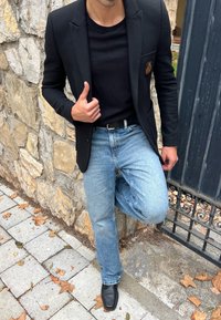 Černý blazer se zlatým emblémem, spárovaný s černým tričkem a světle modrými džíny. Černé kožené boty, na pozadí kamenné zdi.