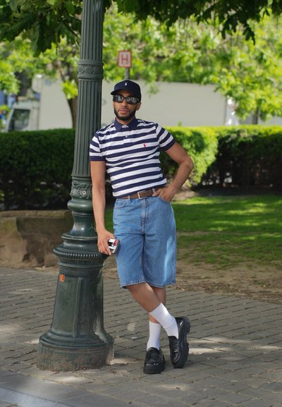 Camisa de polo a rayas azul marino y blanca, pantalones cortos de mezclilla azules, calcetines blancos, zapatos negros y gafas de sol. Apoyado contra un farola verde.