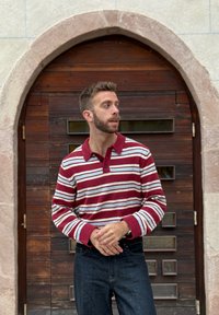 Burgundi ja valge triibuline pullover laia kraega ja kolme nupuga, seljas tumelad sinised teksad; seisab puidust ukse ees.