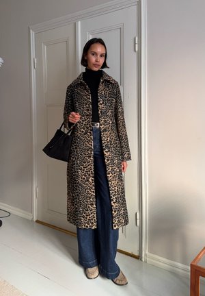 Mujer con abrigo de estampado de leopardo, suéter de cuello alto negro, jeans de pierna ancha, zapatos beige, sosteniendo un bolso negro, de pie en interiores junto a puertas blancas.