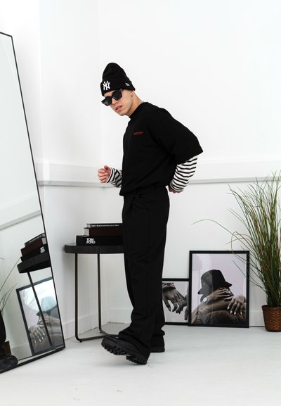 Camiseta negra de manga corta superpuesta sobre una camiseta de rayas de manga larga, pantalones negros, gorro negro, gafas de sol; espejo reflectante y planta de fondo.