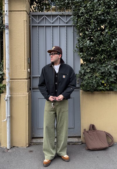 Hombre con chaqueta negra, pantalones anchos verdes, zapatos marrones y gorra, de pie junto a una puerta metálica gris y una pared beige con plantas verdes cercanas.