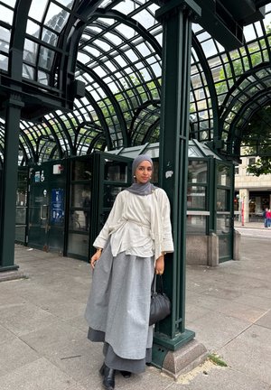 Blusa suelta de color gris claro con diseño drapeado, cinturón plateado y falda amplia gris; bolso negro y botas de tobillo; de pie en una estructura verde arqueada.