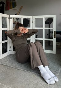 Καφέ μπλούζα με μακριά μανίκια και ταιριαστά παντελόνια με εφαρμοστή σχεδίαση. Λευκές κάλτσες με το κείμενο "TENTH" τυπωμένο πάνω τους. Γκρι υφασμάτινο χαλί πάνω σε πλακάκι.