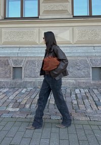 Καφέ δερμάτινο μπουφάν πάνω από μαύρο πουκάμισο, σκούρο τζιν και γκρι σουέτ μπότες. Κρατάει μία καφέ υφασμάτινη τσάντα ενώ περπατάει σε κυβόλιθους.