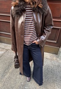 Άτομο που φοράει καφέ δερμάτινο παλτό με γούνινο γιακά, ριγέ πουκάμισο, σκούρα τζιν φαρδύ πόδι και καφέ παπούτσια, κρατώντας μια μικρή καφέ τσάντα.