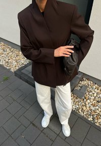 Blazer marrone su misura con colletto alto, abbinato a pantaloni bianchi a gamba larga e sneakers bianchi. Porta una piccola borsa in pelle nera.