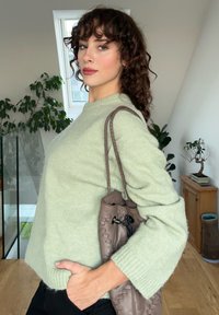 Ανοιχτό πράσινο πουλόβερ με ριχτές μανσέτες, συνδυασμένο με μια καφέ πλεκτή τσάντα χειρός με κλείσιμο κορδόνι. Το φόντο περιλαμβάνει φυτά και έπιπλα.