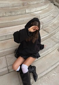 Sweat à capuche noir oversize avec une finition texturée, associé à une jupe noire à volants, des chaussettes blanches montant aux genoux et des bottes noires pointues montant aux genoux.