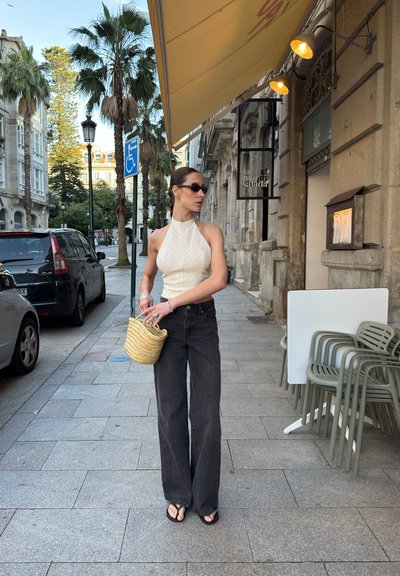 Top halter blanco, jeans de pierna ancha negros, chanclas negras, bolso de paja trenzada. Escenario urbano con palmeras y fachadas de edificios.