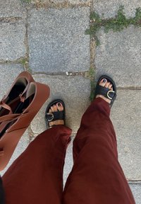 Schwarze Sandalen mit einem breiten Riemen und einer runden Schnalle, kombiniert mit weitgeschnittenen rostfarbenen Hosen und einer braunen Tragetasche, auf steinernem Pflaster.