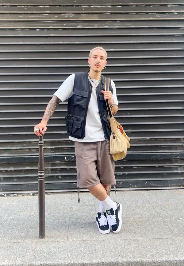 Schwarze ärmellose Weste über einem weißen T-Shirt, braune Shorts, schwarz-weiße Turnschuhe und eine beigen Tasche. Auffällige Tattoos an den Armen.
