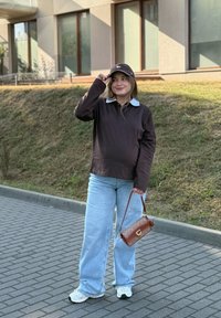 Jonge vrouw buiten in een zwarte pet, een donkerlang mouwshirt, lichtblauwe jeans en witte sneakers, met een bruine handtas in haar hand, staand op een betegeld pad.