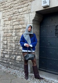 Blue knitted jumper, grey scarf, black handbag, high brown boots. Textured stone wall in the background, black door with metal accents.