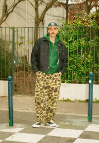 Veste en denim sur un sweat à capuche vert, pantalon à imprimé camouflage, baskets noires. Debout dans une rue avec de la verdure et une clôture en métal en arrière-plan.