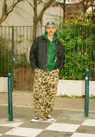 Chaqueta vaquera sobre sudadera con capucha verde, pantalones con estampado de camuflaje, zapatillas negras. De pie en una calle con vegetación y una verja metálica al fondo.