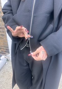 Sweat à capuche noir avec fermeture éclair et poches, porté avec un pantalon noir. Des mains aux ongles longs ornés d'un vernis rouge ajustent la fermeture éclair. Tatouages visibles sur les mains.