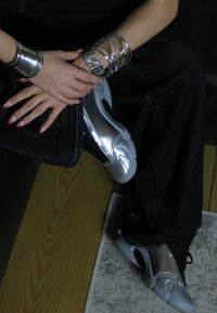 Hopeakuosiset, tekstuuriltaan ballet-tyyliset kengät mustilla nauhoilla ja korostus yksityiskohdilla, paritettu metallisten rannekkeiden kanssa kädessä, joka lepää tummassa farkkukankaassa.