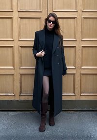 Black double-breasted coat with wide lapels, layered over a black turtleneck and mini skirt, paired with brown knee-high boots and sunglasses.
