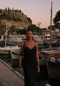 Vestido de seda preto com detalhes em renda, usado por uma pessoa de pé perto de barcos atracados em um porto ao crepúsculo, com uma encosta rochosa ao fundo.
