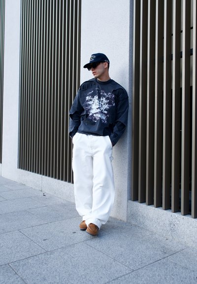 Camisa negra de manga larga con gráfico blanco, pantalones amplios blancos y calzado marrón. De pie contra una pared texturizada con listones verticales.