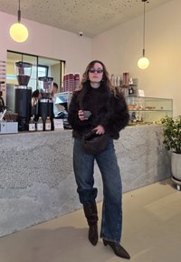 Barna műszőrméből készült kabát, sötét felső, bő, kék farmernadrág, barna, laza csizmák. Egy kávézóban szürke bögrét tart, ahol betonpultok és kávédaráló található.