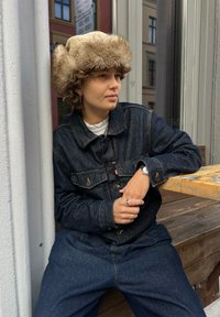 Jeansowa kurtka z metalowymi guzikami, w ciemnoniebieskim kolorze, noszona na białej koszuli. Wełniana czapka z futrzanym wykończeniem. Siedząc na drewnianej ławce, w zrelaksowanej pozycji.
