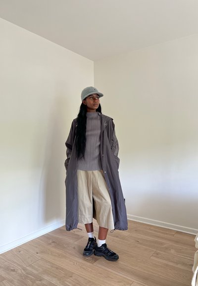 Abrigo largo gris, suéter gris de canalé, pantalones cortos beige y zapatillas negras con calcetines blancos. Los accesorios incluyen una gorra gris claro.