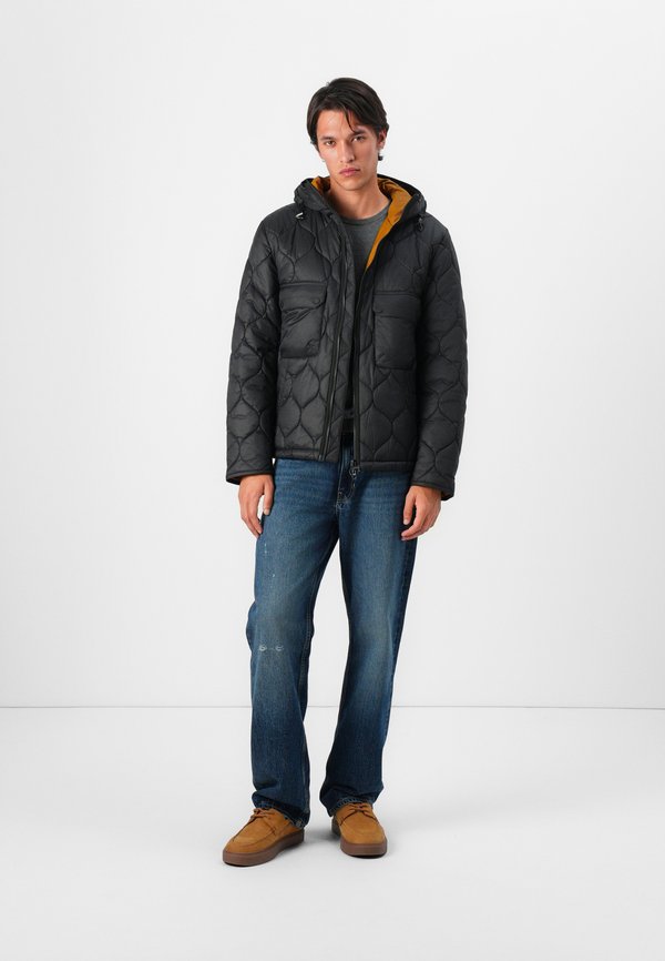 Casaco puff preto acolchoado com capuz, duas bolsos frontais, material leve, usado sobre uma camisa cinza, combinado com jeans azuis e sapatos castanhos.