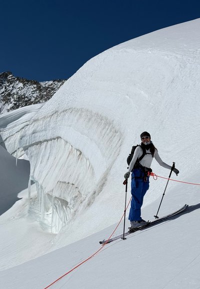 Esquiador con pantalones azules y camiseta blanca de pie en una pendiente nevada cerca de una gran cornisas de nieve, asegurado con cuerda para seguridad contra un telón de fondo de montaña helada.
