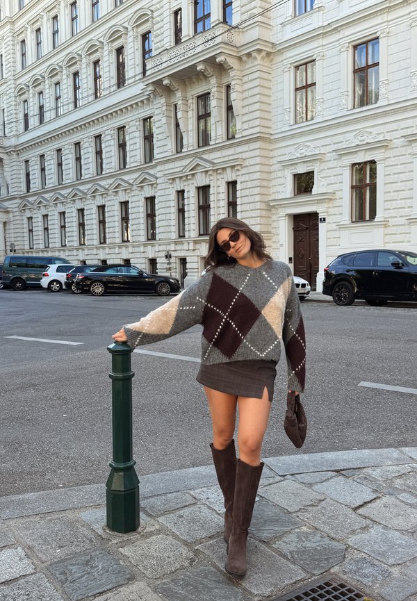 Mujer con gafas de sol, un suéter de rombos, minifalda y botas hasta la rodilla se apoya en un poste verde en una calle de la ciudad con edificios blancos históricos.