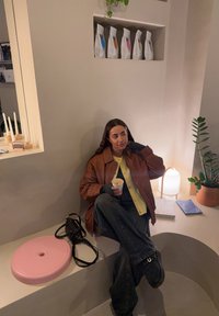 Femme en veste marron assise dans un coin beige incurvé tenant une tasse, avec un sac noir, un siège rose, une plante, une lampe et des produits emballés sur l'étagère murale.