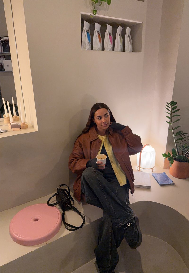 Femme en veste marron assise dans un coin beige incurvé tenant une tasse, avec un sac noir, un siège rose, une plante, une lampe et des produits emballés sur l'étagère murale.