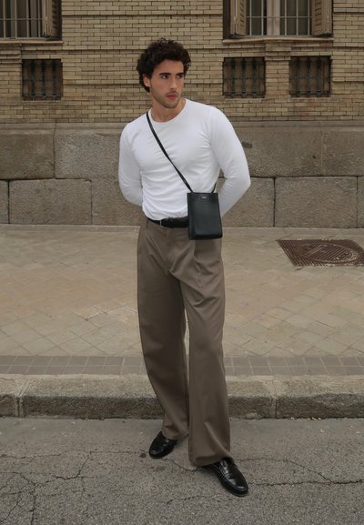 Camisa blanca de manga larga, pantalones anchos beige, cinturón negro y bolso cruzado negro. Lleva zapatos de cuero negros. De pie en una calle pavimentada.