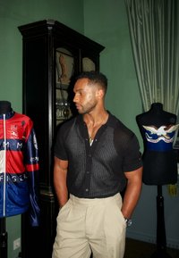 Svart kortärmad stickad skjorta med öppen väv, beige byxor och silverfärgad klocka. Man står bredvid en röd och blå jacka och en mannequin.