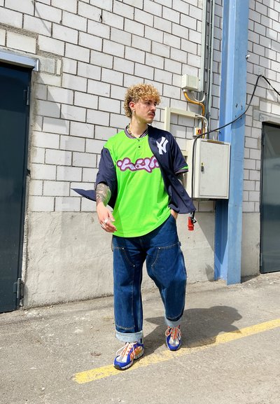 Jersey de malla verde con mangas azul marino, que presenta letras en blanco y púrpura. Combinado con jeans de mezclilla holgados y zapatillas multicolores.