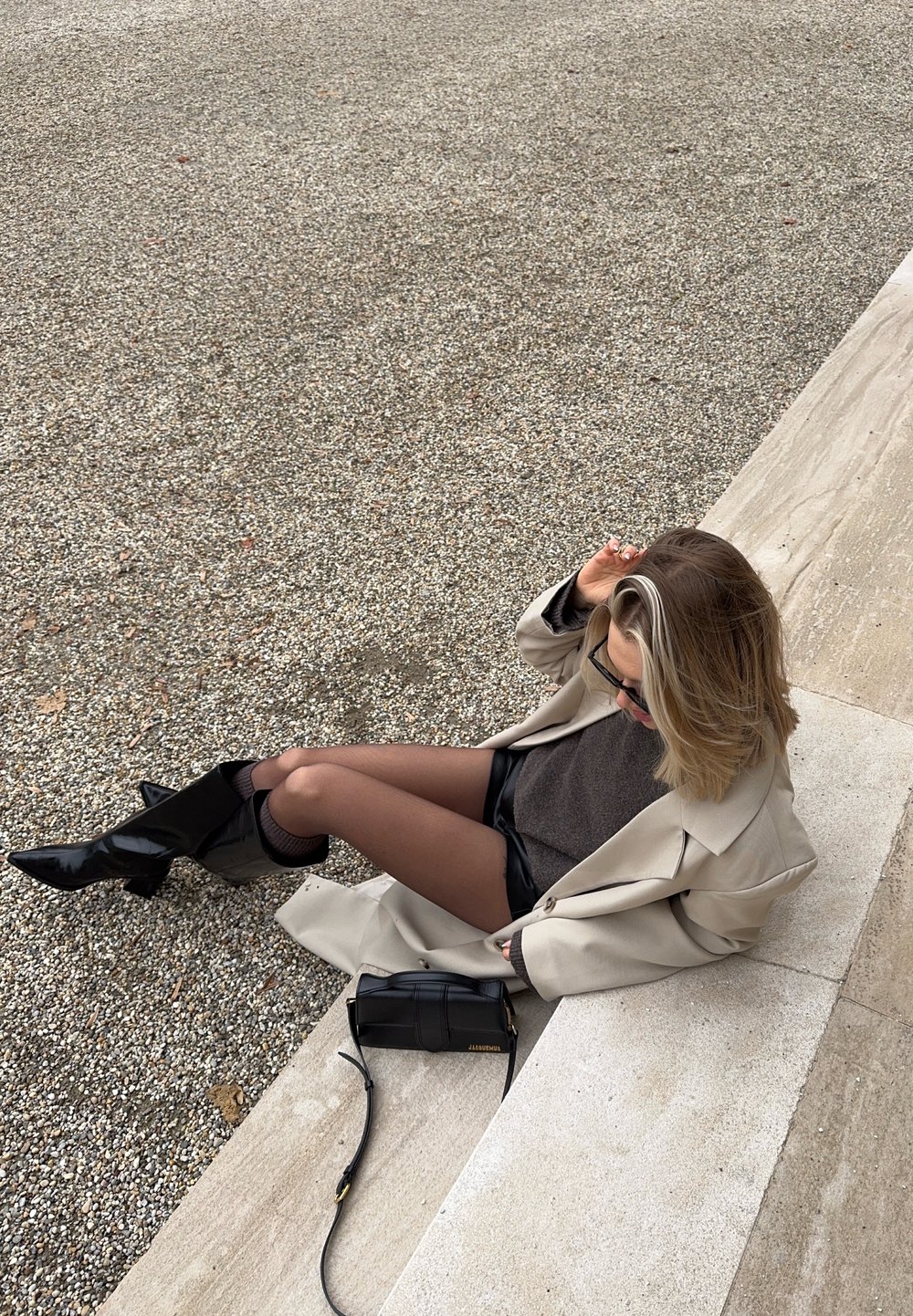 Bézs túlméretezett kabát sötét pulóver fölött és fekete rövidnadrággal; fényes fekete térdig érő csizmák; kis fekete kézitáska a földön.