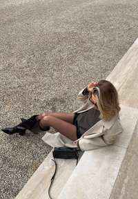 Bézs túlméretezett kabát sötét pulóver fölött és fekete rövidnadrággal; fényes fekete térdig érő csizmák; kis fekete kézitáska a földön.