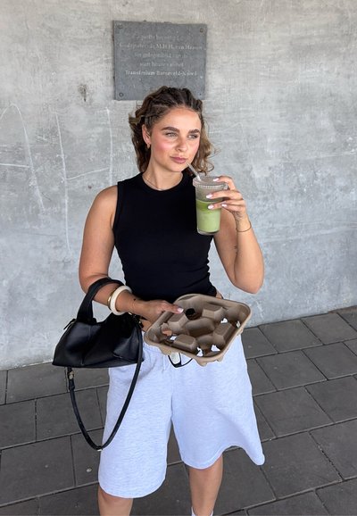 Top sin mangas negro, pantalones cortos grises, sosteniendo una taza transparente con una bebida verde y un portador de cartón marrón. Bolso negro con correa para el hombro.