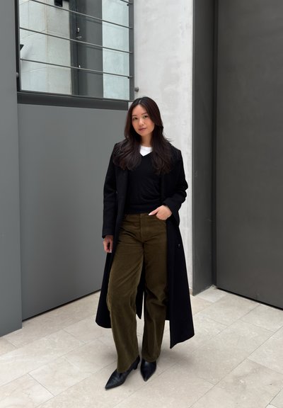 Mujer de pie en el interior vistiendo un abrigo negro largo, camiseta negra, pantalones verde oliva y zapatos negros puntiagudos frente a paredes grises y suelo de baldosas.