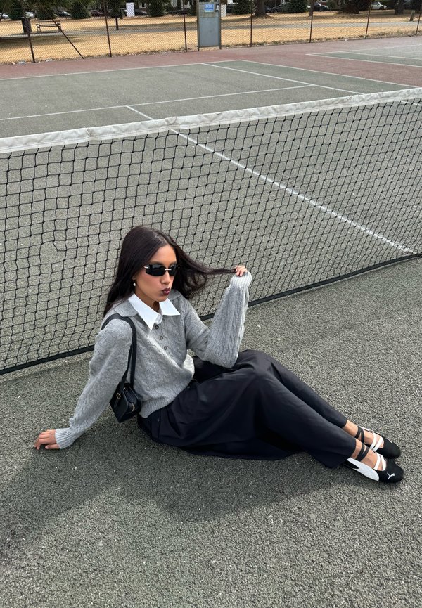 Donna con maglione grigio, gonna nera e occhiali da sole seduta su un campo da tennis vicino alla rete, tenendo i capelli con una mano.