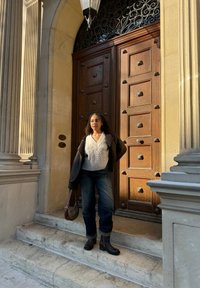 Porte en bois brun avec des motifs carrés ; le modèle porte un pull en mailles blanches, un jean sombre et des bottines noires, debout sur des marches en pierre.
