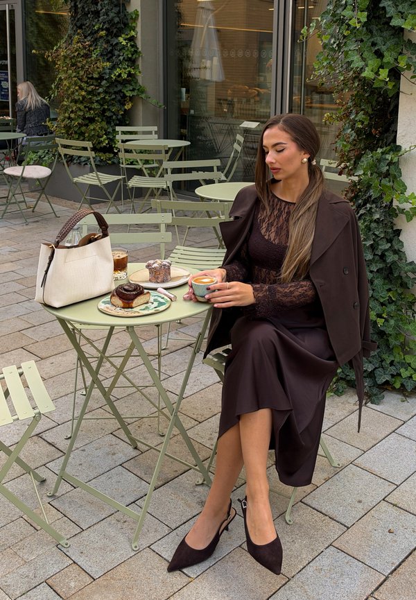 Una mujer está sentada a la mesa de una cafetería al aire libre, con café, bollería y un bolso, rodeada de sillas verdes y ventanas cubiertas de hiedra.