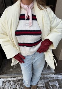 Άτομο με εκρού μπουφάν puffer, ριγέ πλεκτό πουλόβερ, ανοιχτό μπλε τζιν, ροζ φουλάρι λαιμού, κόκκινα δερμάτινα γάντια, στέκεται σε χιονισμένο έδαφος.