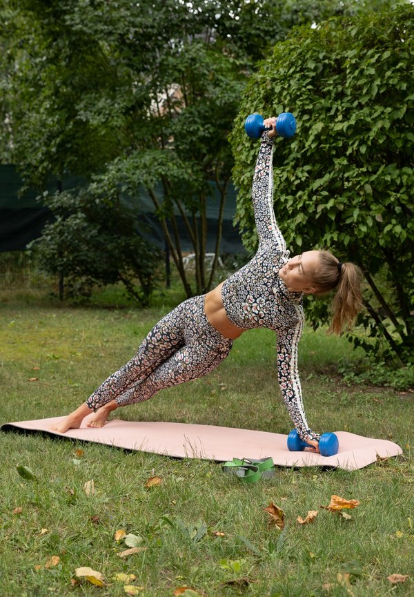 Una mujer mantiene una plancha lateral sobre una esterilla al aire libre, apoyándose en una mano y levantando una mancuerna azul con la otra.