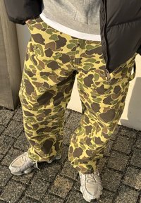 Personne portant un pantalon camouflage jaune et vert, des baskets gris clair, une veste noire et un sweat à capuche gris, debout sur une surface pavée.